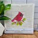 A packaged needlepoint kit showing a cardinal bird with green holly leaves on a white mesh canvas, with a branded label at the bottom reading Blue Poppy Designs Needlepoint Kit. The package is photographed on a wooden surface with a plant and white tile wall in the background.