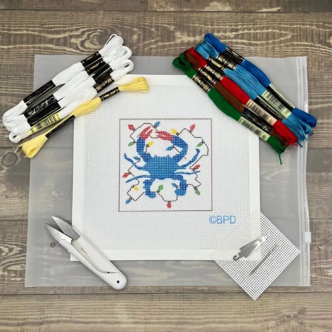 A cross-stitch kit laid out on a wood surface, including a white A4 sheet with a colorful crab design, multiple skeins of thread in white, blue, red, green, and yellow, scissors, a utility knife, and a small metal tool.