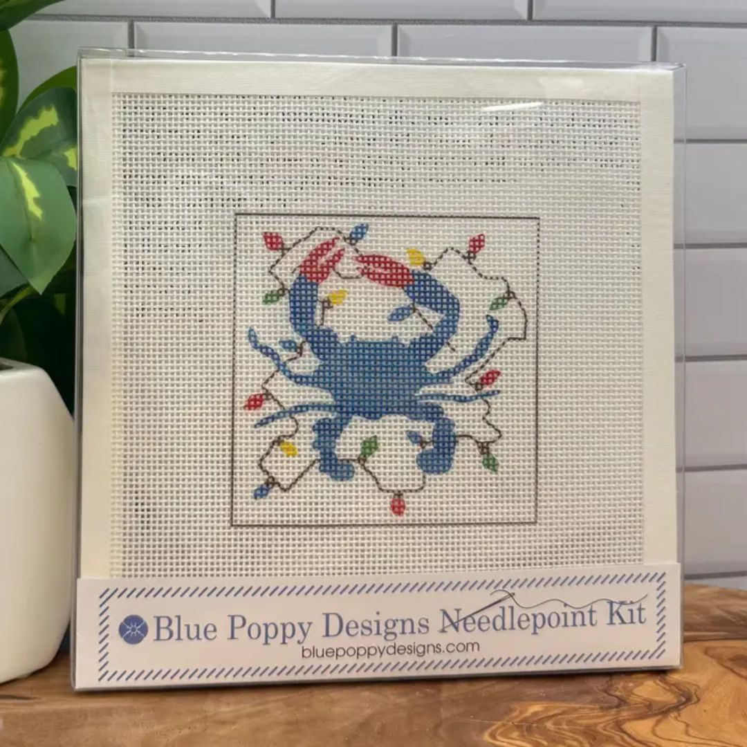 A needlepoint kit in its clear plastic sleeve, featuring a blue crab design with colorful Christmas lights on a white mesh canvas. The packaging label at the bottom reads 'Blue Poppy Designs Needlepoint Kit'.