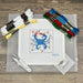 A cross-stitch kit laid out on a wood surface, including a white A4 sheet with a colorful crab design, multiple skeins of thread in white, blue, red, green, and yellow, scissors, a utility knife, and a small metal tool.