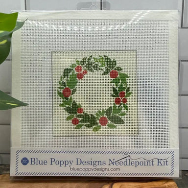 Blue Poppy Designs Needlepoint Kit featuring a festive wreath design with green leaves and red berries on a white mesh canvas, packaged in a clear plastic sleeve with a branded label at the bottom.