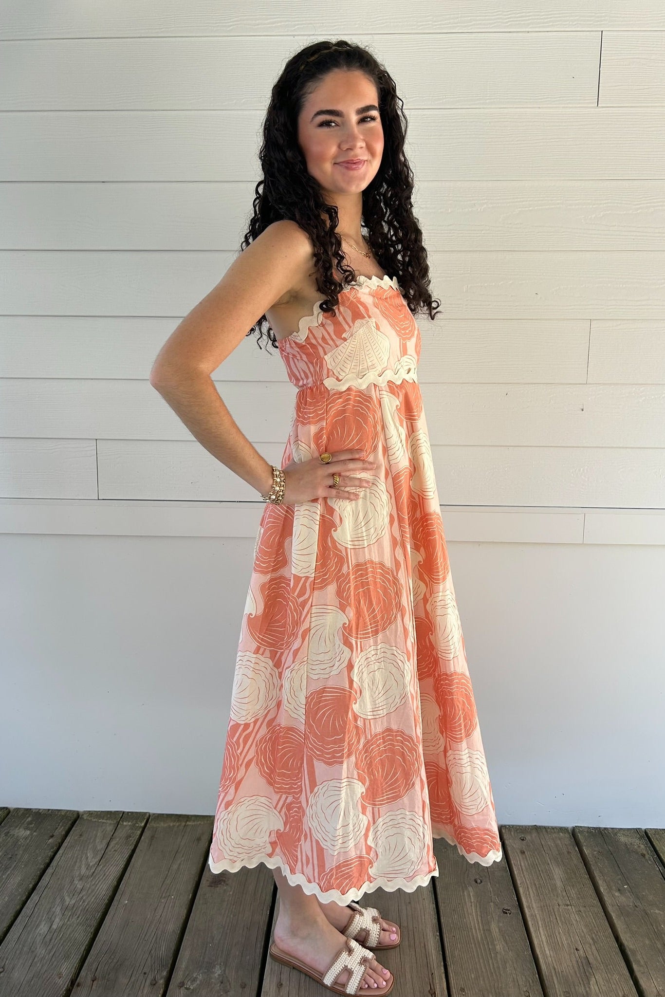 Woman wearing a seashell patterned dress standing on a wooden deck with a white wall background