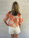 Person wearing a floral blouse and white shorts against a light wooden panel background