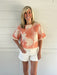 Woman wearing a coral floral blouse and white shorts against a white wooden background