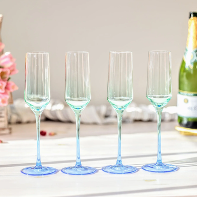 Four clear champagne flutes with a light blue gradient at the base, arranged in a row on a white surface with a blurred champagne bottle in the background.