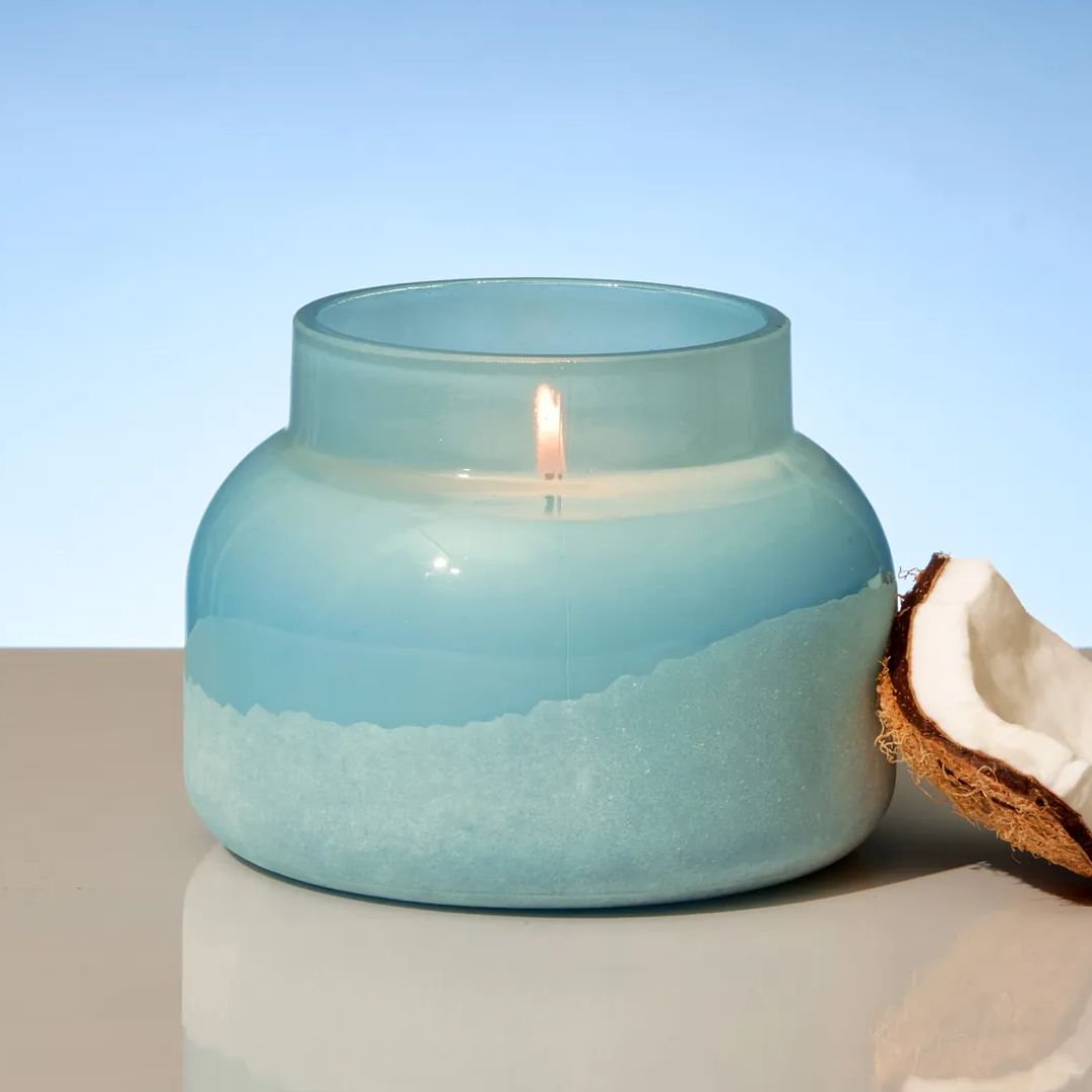 A light blue glass jar candle with a wick and flame, styled next to a halved coconut on a neutral surface against a soft blue gradient background.