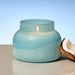 A light blue glass jar candle with a wick and flame, styled next to a halved coconut on a neutral surface against a soft blue gradient background.