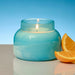 A lit teal-blue glass jar candle sits on a reflective surface with orange slices beside it against a blue gradient background.