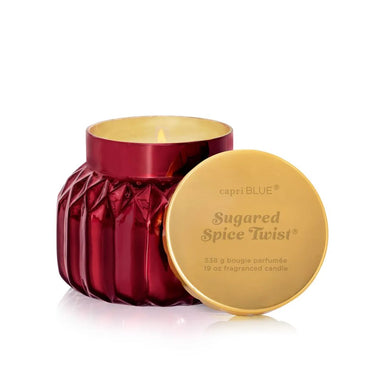 A red faceted glass jar candle with its gold lid leaning against it on a white background. The lid is printed with the brand name 'capri BLUE' and the scent name 'Sugared Spice Twist'.