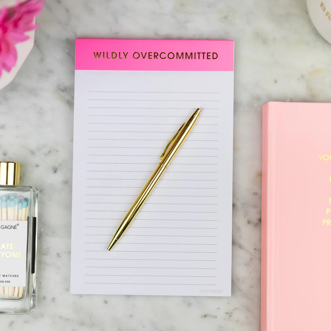 Notepad with 'Wildly Overcommitted' text, gold pen, and perfume bottle on a marble surface