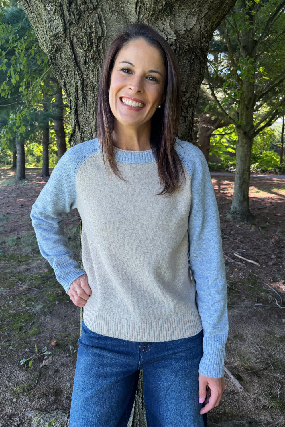 A woman smiling outdoors in front of a tree, wearing a light heather gray crewneck sweater with light blue raglan sleeves and blue jeans.