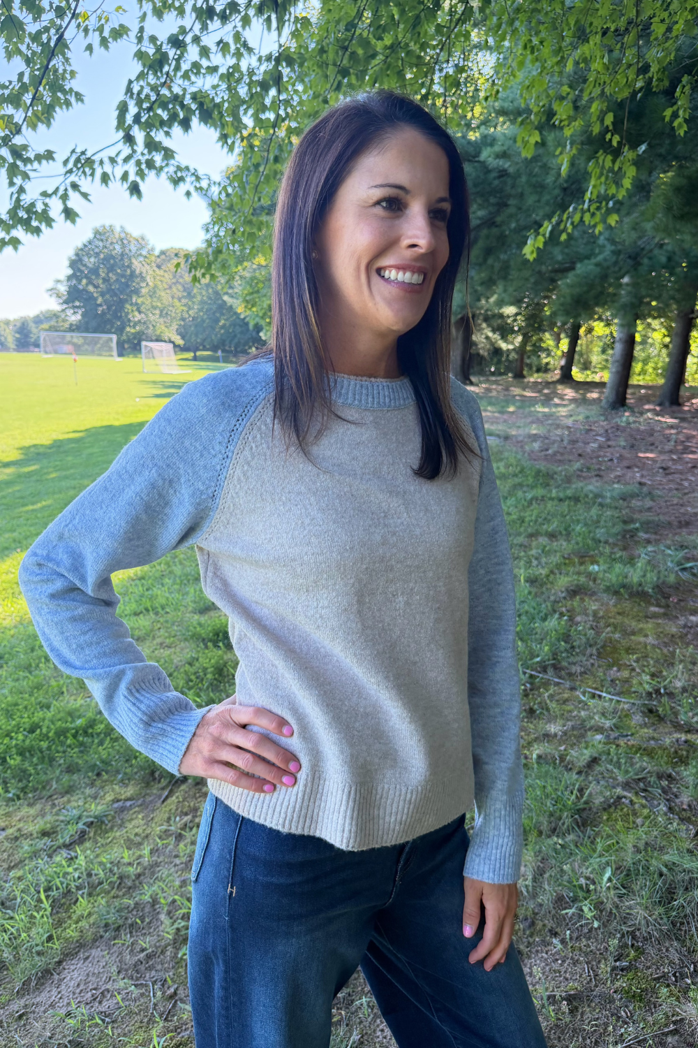 A woman wearing a light gray and blue raglan sweater with dark blue jeans, standing outdoors in a grassy park area with trees.