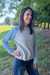 A woman wearing a light gray and blue raglan sweater with dark blue jeans, standing outdoors in a grassy park area with trees.