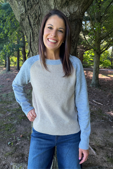 A woman smiling outdoors in front of a tree, wearing a light heather gray crewneck sweater with light blue raglan sleeves and blue jeans.