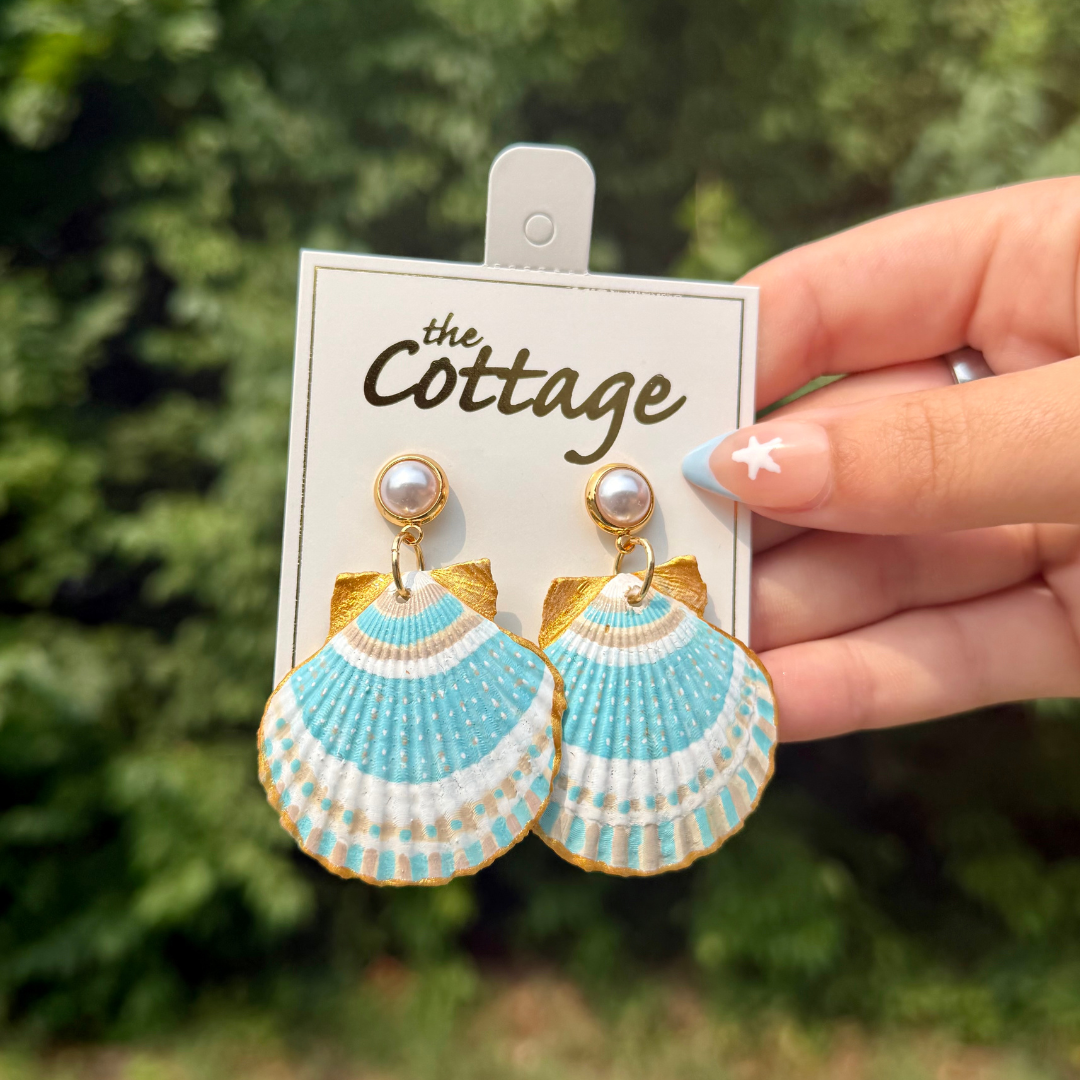 A hand holds a white display card featuring a pair of shell-shaped drop earrings. The earrings have a light blue and white striped pattern with gold trim, gold-tone posts, and a small white pearl at the top.