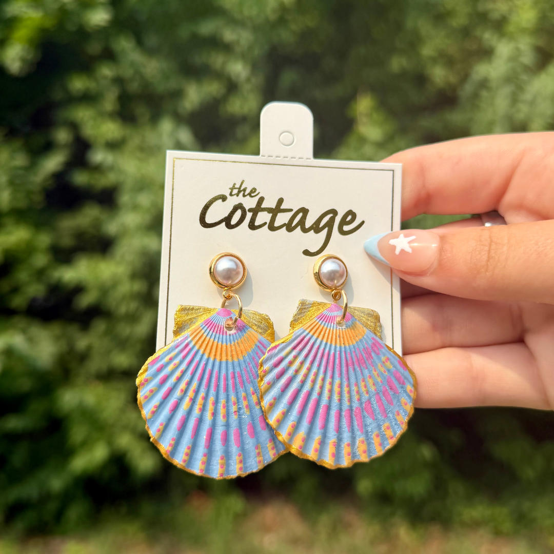 A hand holds a pair of colorful shell-shaped drop earrings on a white display card labeled 'The Cottage.' The earrings have a scallop pattern in pastel blue, pink, purple, and yellow with gold-tone metal and pearl-like studs.