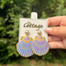 A hand holds a pair of colorful shell-shaped drop earrings on a white display card labeled 'The Cottage.' The earrings have a scallop pattern in pastel blue, pink, purple, and yellow with gold-tone metal and pearl-like studs.
