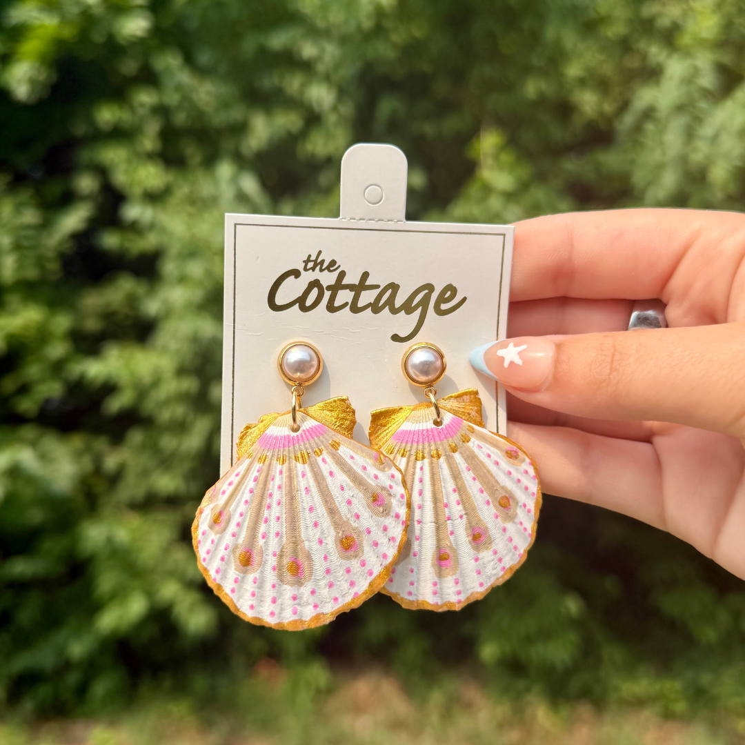 A pair of shell-shaped dangle earrings with gold-tone metal, pink and white embroidery, and pearl studs, displayed on a white card labeled 'The Cottage' held by a hand against a blurred green outdoor background.