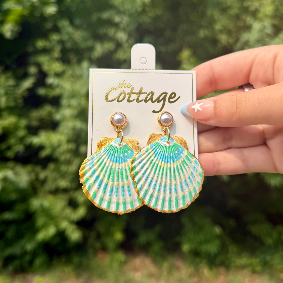 A hand holds a pair of scallop shell drop earrings with a green, blue, and white pattern and gold trim, attached to a white display card from 'The Cottage', against a blurred green outdoor background.