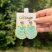 A hand holds a pair of scallop shell drop earrings with a green, blue, and white pattern and gold trim, attached to a white display card from 'The Cottage', against a blurred green outdoor background.
