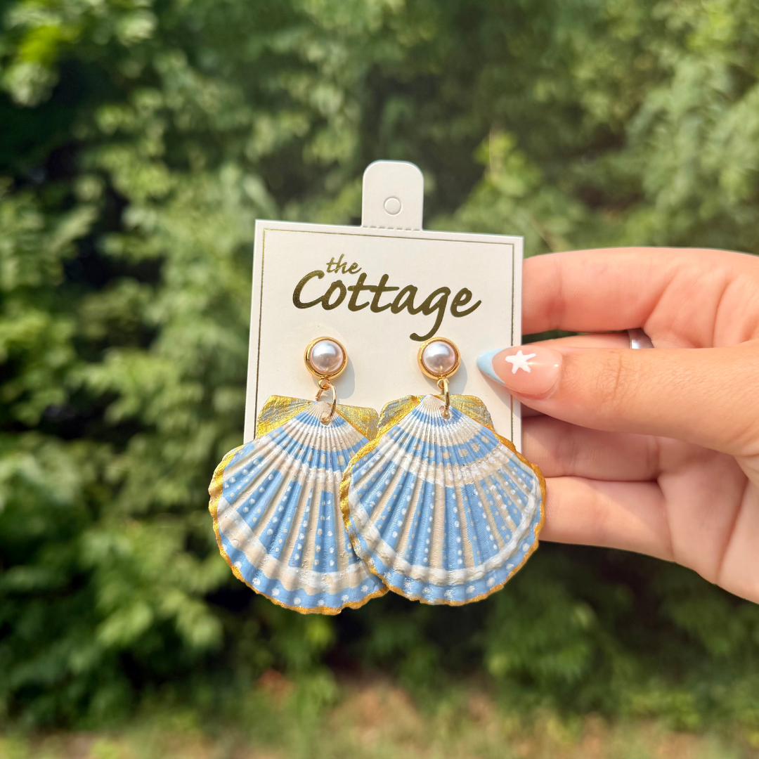 A hand holds a pair of shell-shaped dangle earrings on a display card. The earrings are light blue and white patterned with gold trim, and each has a small pearl-like bead at the top.