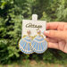 A hand holds a pair of shell-shaped dangle earrings on a display card. The earrings are light blue and white patterned with gold trim, and each has a small pearl-like bead at the top.