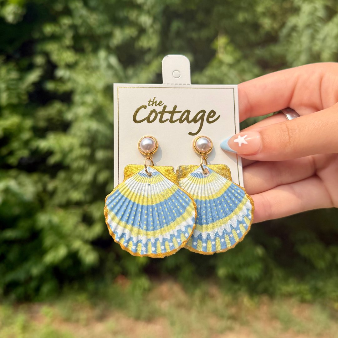 A hand holds a pair of shell-shaped dangle earrings on a display card labeled 'The Cottage.' The earrings have a blue, yellow, and white scallop pattern with gold-tone posts and pearl-like studs.
