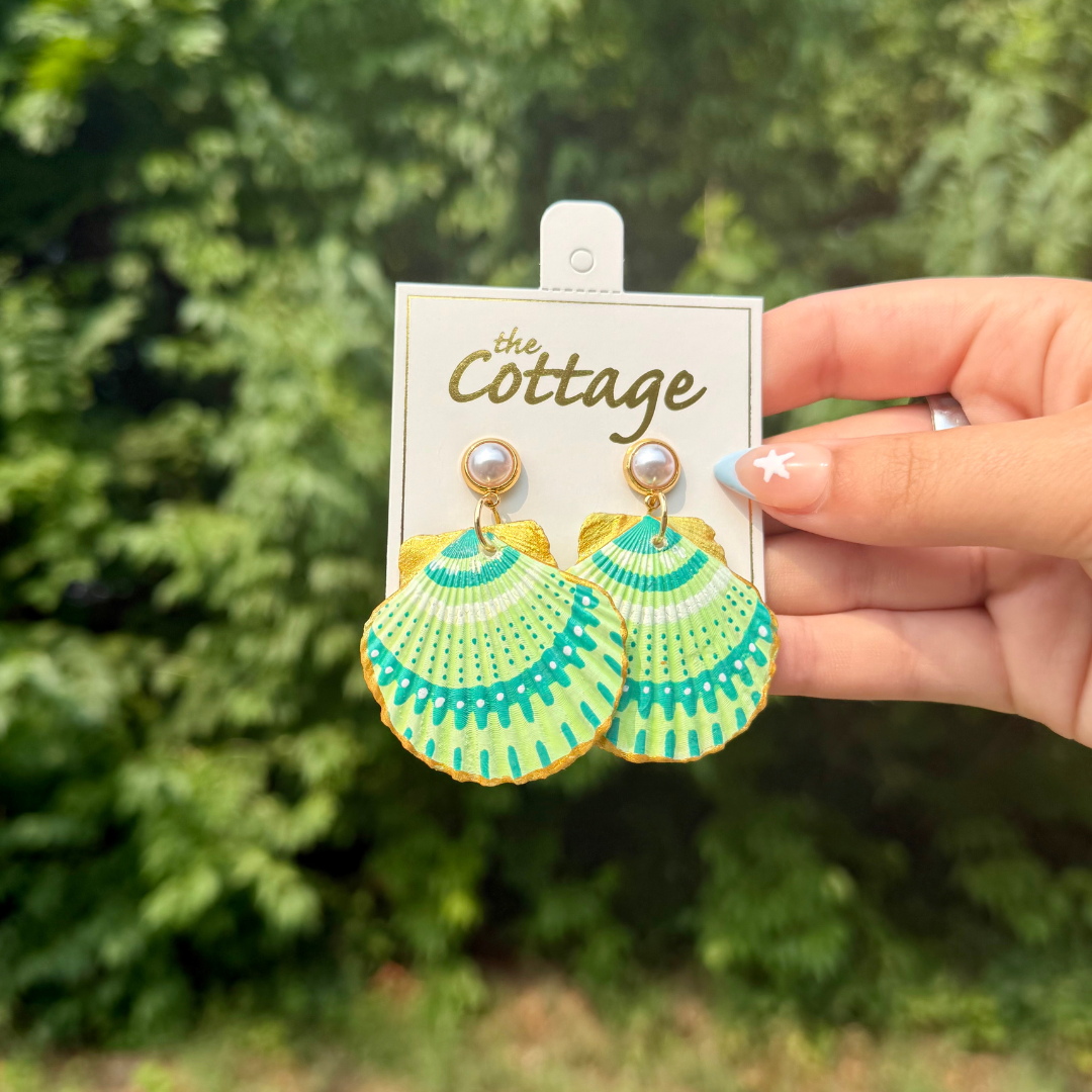 A hand holds a pair of shell-shaped dangle earrings with turquoise and green striped patterns and gold-tone details, attached to a white display card labeled 'the Cottage' against a blurred green outdoor background.