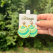 A hand holds a pair of shell-shaped dangle earrings with turquoise and green striped patterns and gold-tone details, attached to a white display card labeled 'the Cottage' against a blurred green outdoor background.