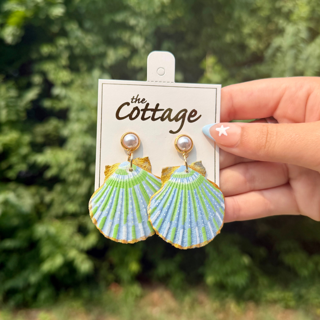 A hand holds a pair of shell-shaped drop earrings on a white display card labeled 'The Cottage.' The earrings have a scallop shell design in light blue and green with gold-tone edges and pearl studs at the top.