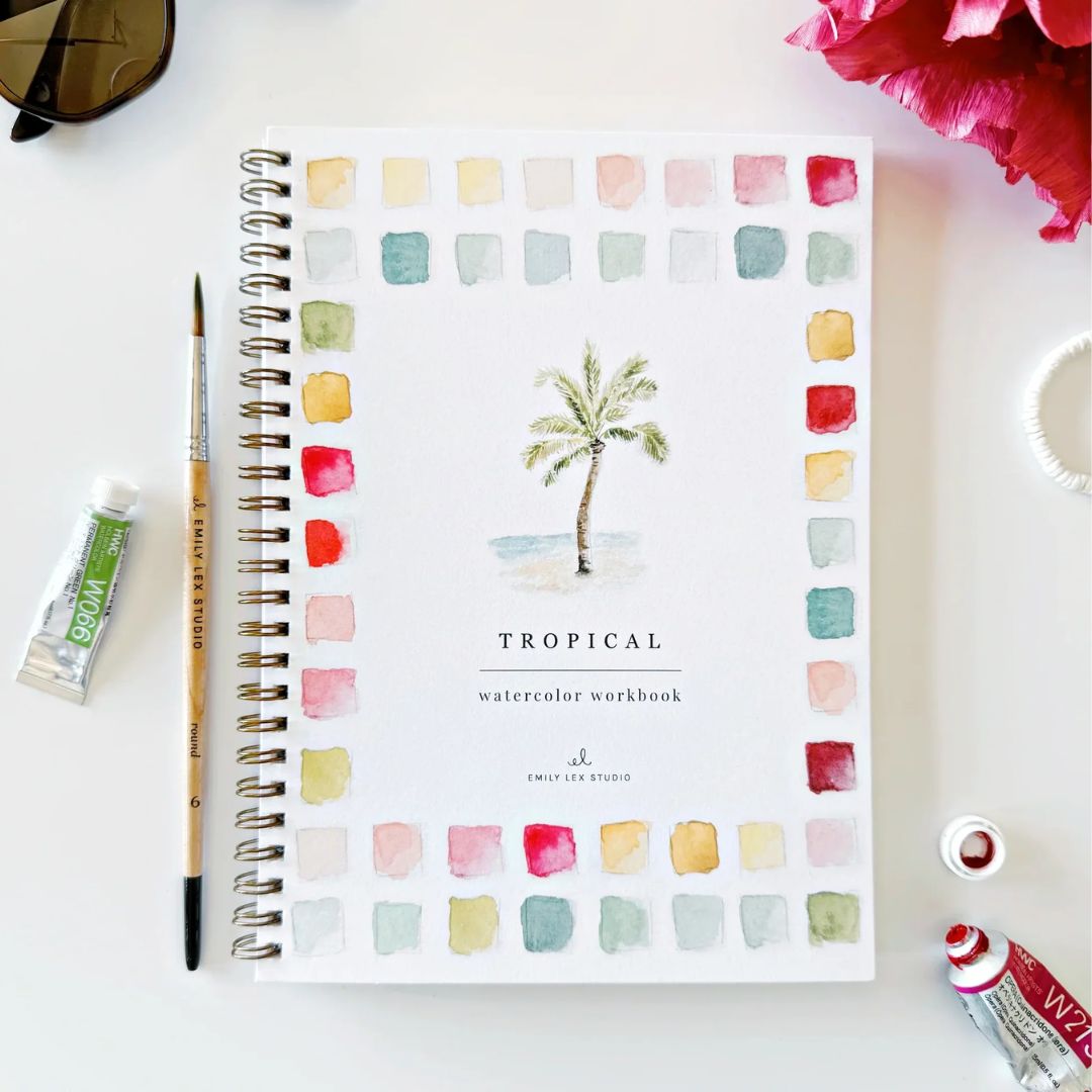 Tropical watercolor workbook with a painted palm tree and colorful watercolor swatches on the cover, styled on a white surface with a paintbrush, watercolor paint tubes, sunglasses and bright pink flowers.