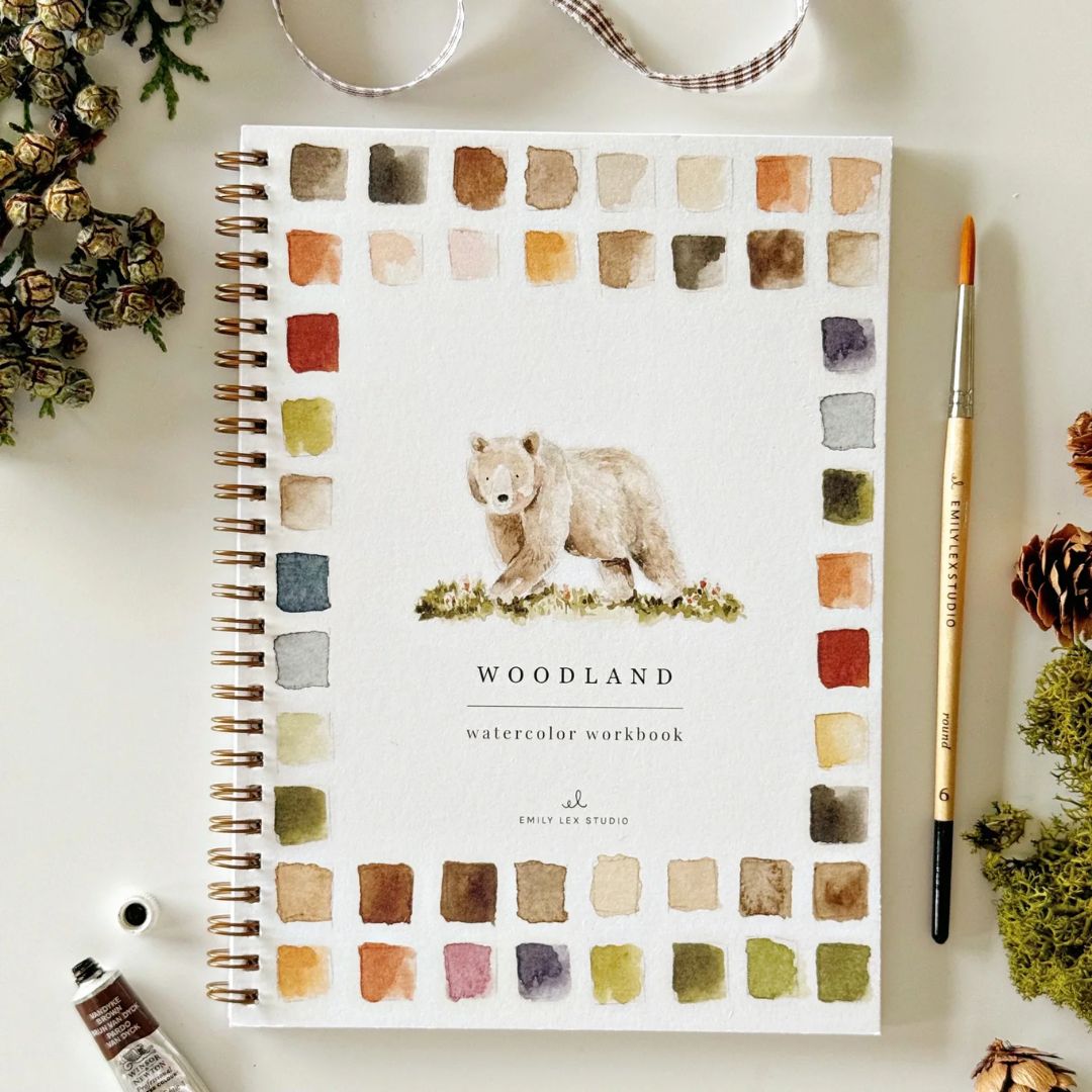 Woodland watercolor workbook featuring a painted bear and earthy watercolor swatches, styled on a light surface with a paintbrush, watercolor paint, dried flowers, pinecones and greenery.