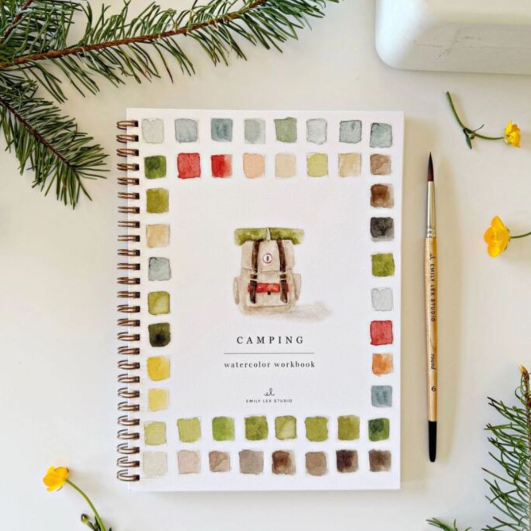Camping watercolor workbook featuring a painted backpack surrounded by colorful watercolor swatches, styled with a paintbrush, evergreen branches and small yellow flowers on a light surface.