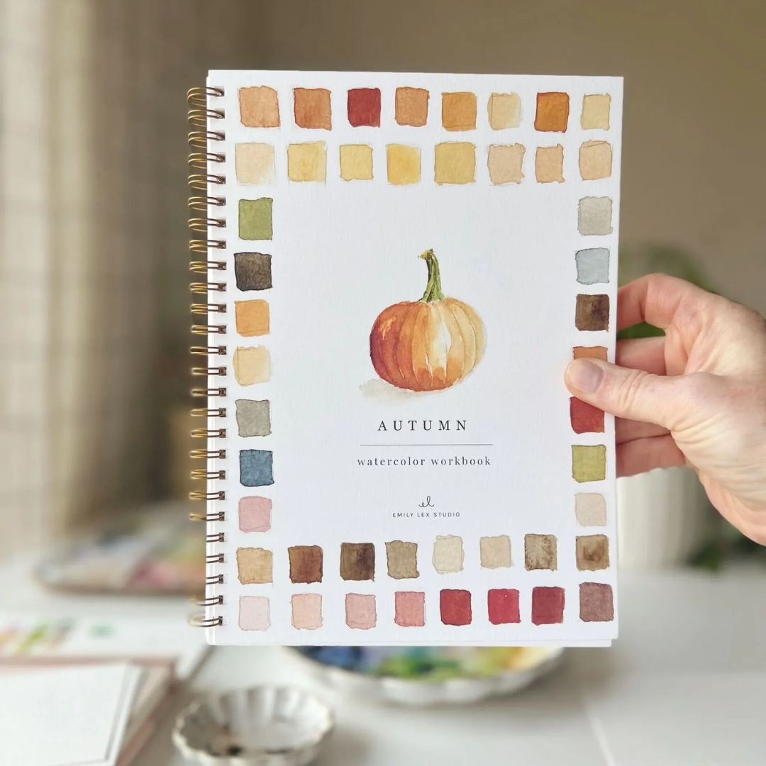 Autumn watercolor workbook featuring a painted orange pumpkin and fall-colored watercolor swatches, held by a hand with a softly blurred art workspace in the background.