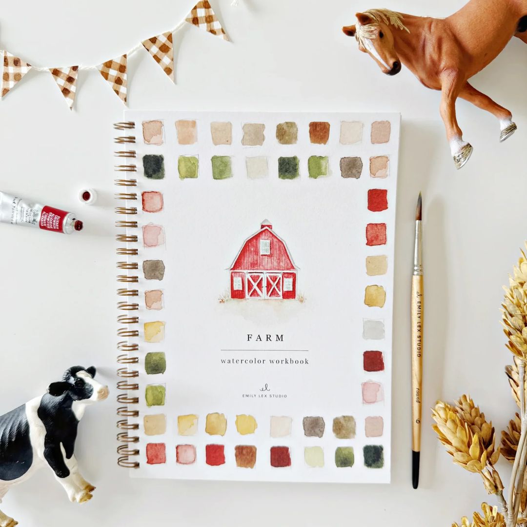 Farm watercolor workbook featuring a painted red barn and colorful watercolor swatches, styled with a paintbrush, watercolor paint, toy cow and horse, checkered bunting and dried wheat on a white surface.