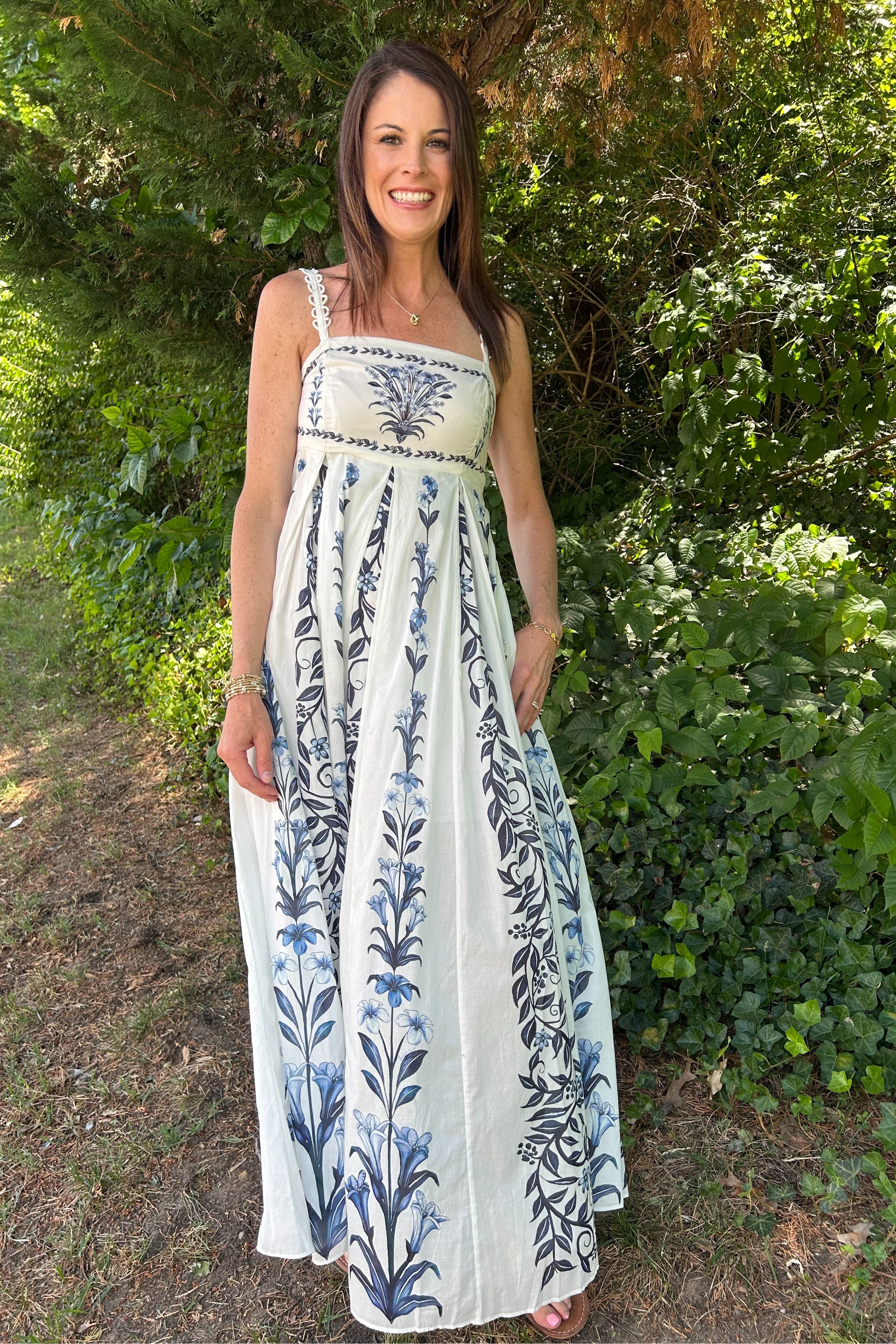 Woman wearing a white dress with blue floral patterns standing outdoors among greenery.