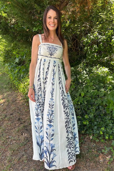 Woman wearing a white dress with blue floral patterns standing outdoors among greenery.