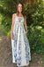 Woman wearing a white dress with blue floral patterns standing outdoors among greenery.