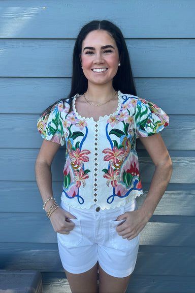 Woman wearing a colorful embroidered blouse and white shorts standing against a blue wall.