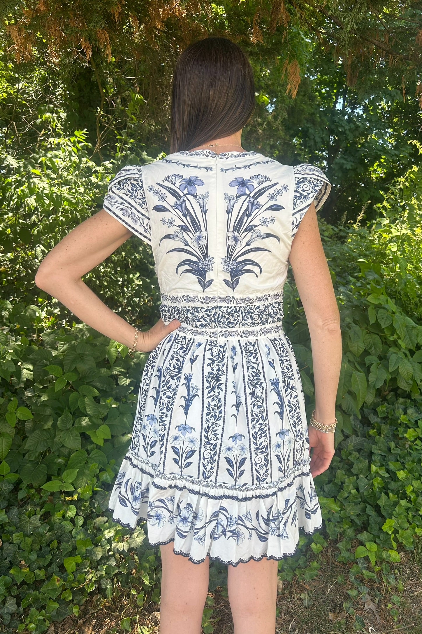 Person wearing a white dress with blue floral patterns standing in front of green foliage.