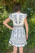 Person wearing a white dress with blue floral patterns standing in front of green foliage.