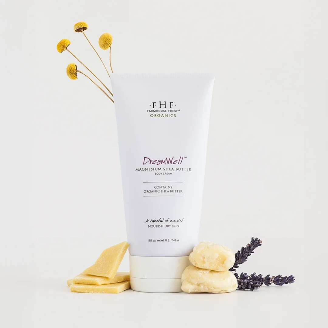 A white tube of FHF Farmhouse Fresh Organics DreamWell Magnesium Shea Butter body cream, styled with yellow flowers, soap bars, and lavender on a clean white background.