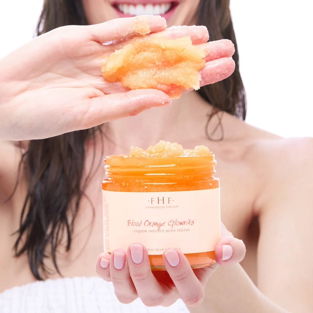 A woman holds an open jar of FHF Face & Body Scrub in the Blood Orange Glow variant, showing the orange scrub being smeared onto her hand against a clean white background.