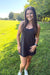 A woman standing outdoors on a grassy field wearing a black sleeveless tank top dress and white sneakers.
