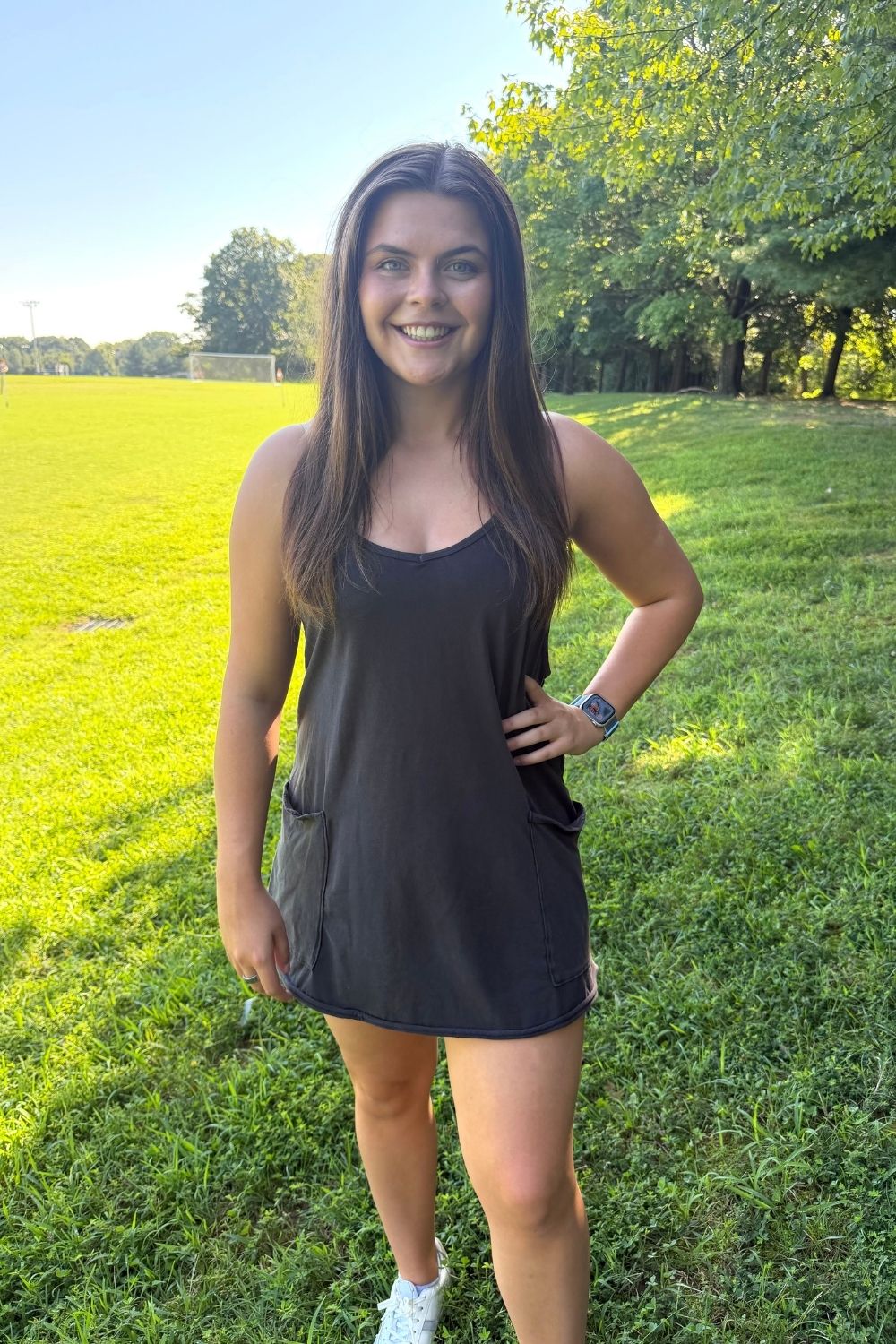 A woman wearing a dark charcoal gray sleeveless tank dress stands on a grassy park lawn with trees in the background.