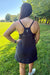 A woman viewed from behind wearing a black athletic tank dress with thin straps, a small silver emblem at the back neckline, and a zippered pocket, standing on a grassy field with trees in the background.