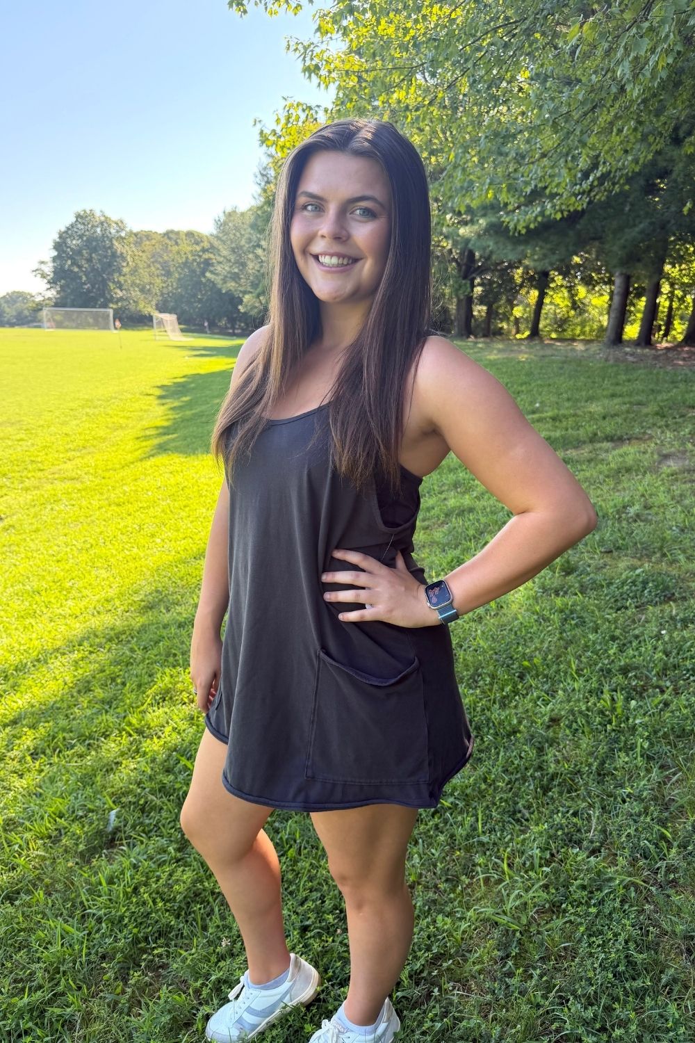A woman posing outdoors in a grassy park wearing a dark charcoal gray sleeveless tankini-style mini dress with a side pocket, paired with white sneakers.