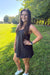 A woman posing outdoors in a grassy park wearing a dark charcoal gray sleeveless tankini-style mini dress with a side pocket, paired with white sneakers.