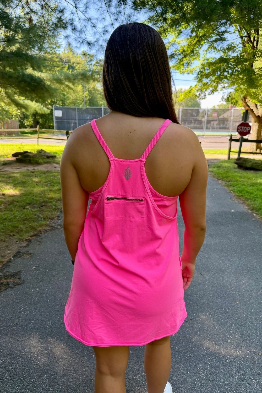 A woman viewed from behind wearing a bright pink athletic tank dress with thin straps and a small zippered pocket on the back, standing on an outdoor paved path with trees and a tennis court in the background.