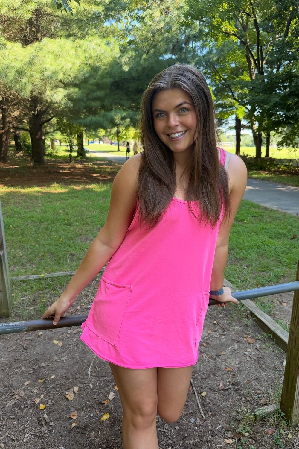 A woman wearing a bright pink sleeveless mini dress with pockets, standing outdoors in a park setting.